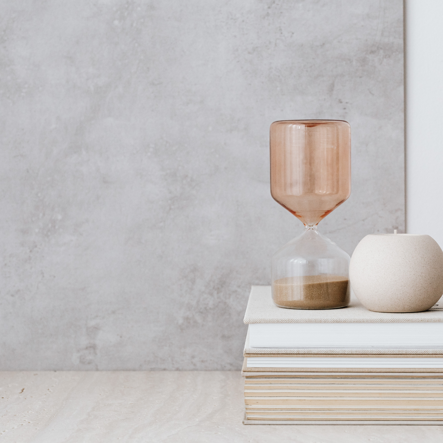 Minimalist hourglass timer and candle on stacked books against a modern concrete wall – curated home accessories for calming interiors